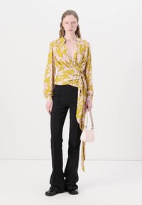 Gelbe Bluse mit floralem Wrap-Design und langen Ärmeln, kombiniert mit schwarzen Schlaghosen. Das Model hält eine kleine rosa Handtasche. Glatte Stofftextur.