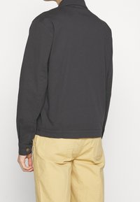 Czarna szara kurtka o luźnym kroju, z kołnierzem, zapinanymi mankietami i bocznymi rozcięciami, w połączeniu z beżowymi spodniami z teksturowanego materiału.