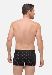 Hombre con la espalda desnuda que lleva puestos bóxers negros con un patrón de pequeñas bicicletas rojas, de pie contra un fondo blanco liso.