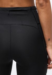 Κοντινό πλάνο μαύρου αθλητικού κολάν με μια μικρή τσεπούλα με φερμουάρ στη μέση, που φοριέται από ένα άτομο με μικρό τατουάζ στον καρπό.