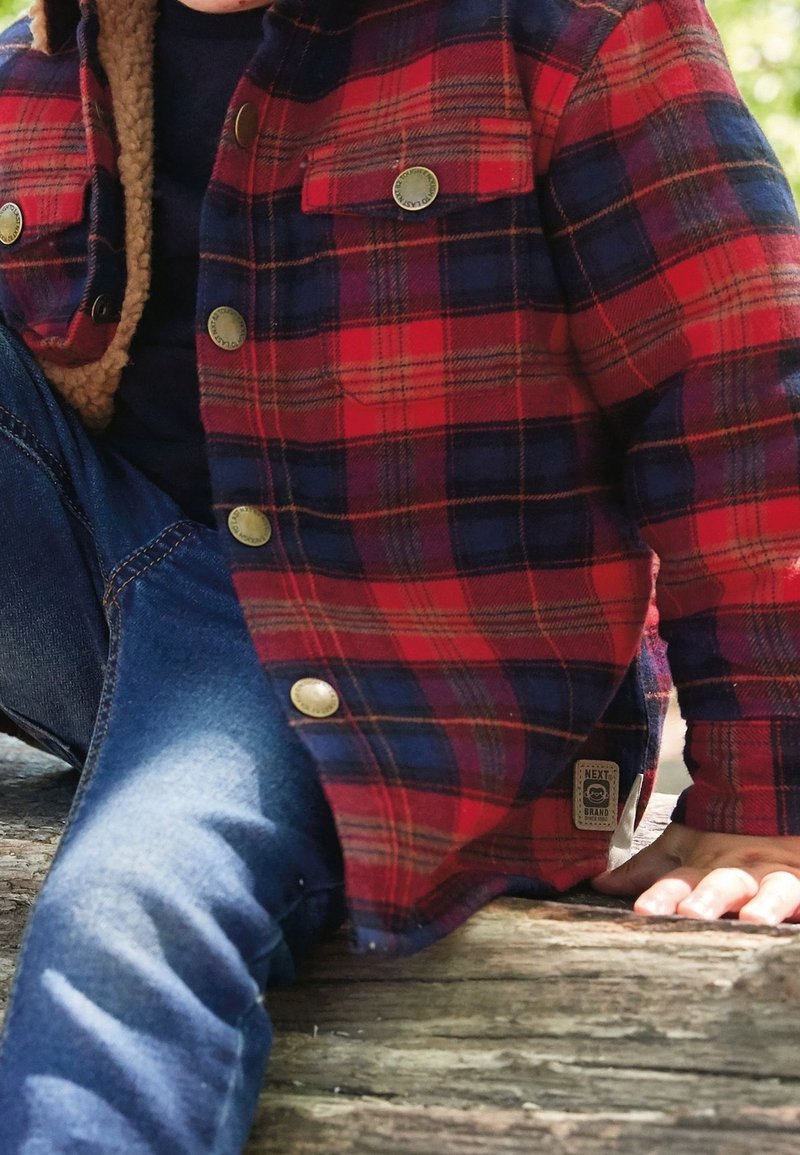 Enfant portant une veste à carreaux rouge et bleu marine avec une doublure beige et un jean bleu, assis sur une surface en bois à l'extérieur, main posée sur le bois.