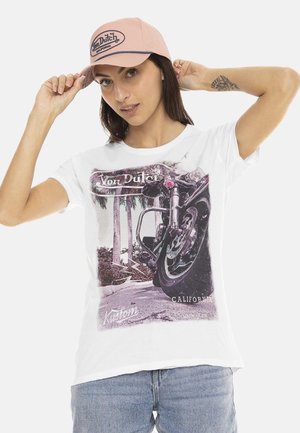Femme portant un t-shirt blanc avec un motif de moto et une casquette de baseball rose Von Dutch, ajustant la casquette avec ses deux mains.