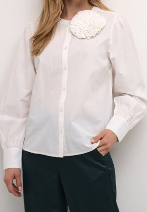 Kvinde iført hvid skjorte med knapper og puffede ærmer samt stor stofblomst på skulderen, kombineret med mørke bukser.