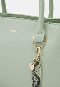 Ljusgrön texturerad handväska med guldfärgad hårdvara, som har ett flätat band i ett ormskinnsmönster. Logotypen är präglad på framsidan.