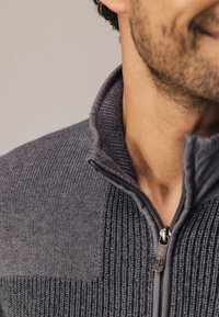 Grijze zip-up sweater met een textuurstof waarin geribbelde en gladde secties zijn verwerkt. Inclusief een hoge kraag en metalen ritsdetail.