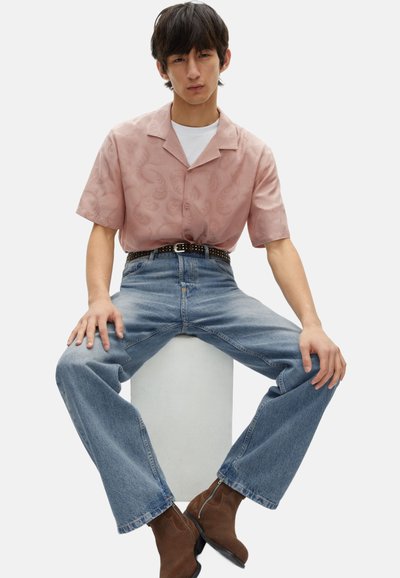 Lichtroze shirt met korte mouwen met een paisleypatroon, gecombineerd met lichtblauwe wijde jeans en bruine enkellaarsjes, zittend op een witte voetstuk.