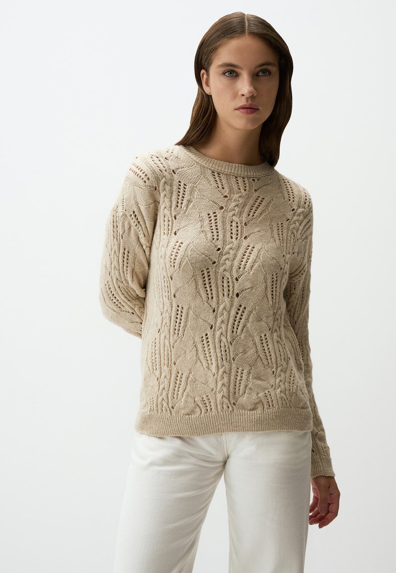 Beiger Strickpullover mit strukturiertem Blattmuster, Rundhalsausschnitt und geripptem Saum. Kombiniert mit hellfarbigen Hosen.