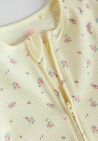 Tissu côtelé crème avec petit motif floral rose et violet, accompagné d'une fermeture éclair centrale partiellement ouverte près d'un col arrondi.