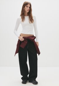 Bílý dlouhý rukáv těsně přiléhající top v kombinaci s volnými černými džíny. Na pase je uvázaná vínová bunda a model má na sobě černé boty.