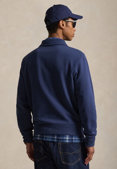 Mand iført navy jakke, blå ternet skjorte, mørke jeans, navy baseballkasket og sorte solbriller, set bagfra mod en neutral baggrund.