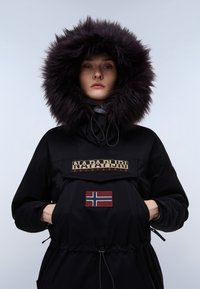 Sort parka med en stor faux fur hætte, der har et iøjnefaldende logo patch og et norsk flag. Inkluderer justerbare snore og lynlåslommer.