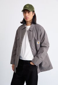 Chaqueta gris con botones a presión y un bolsillo en el pecho, llevada sobre una camisa blanca, combinada con pantalones negros y una gorra verde con el logo de Nueva York.