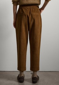 Pantaloni in corduroy marrone con cintura elastica, tasche posteriori, design a gamba affusolata e orli arrotolati, abbinati a scarpe scure e calzini texturizzati.