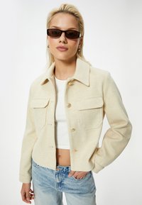 Cremefarvet uldblandingjakke med krave, to forlommer og gyldne knaplukninger, parret med lyseblå denim jeans.
