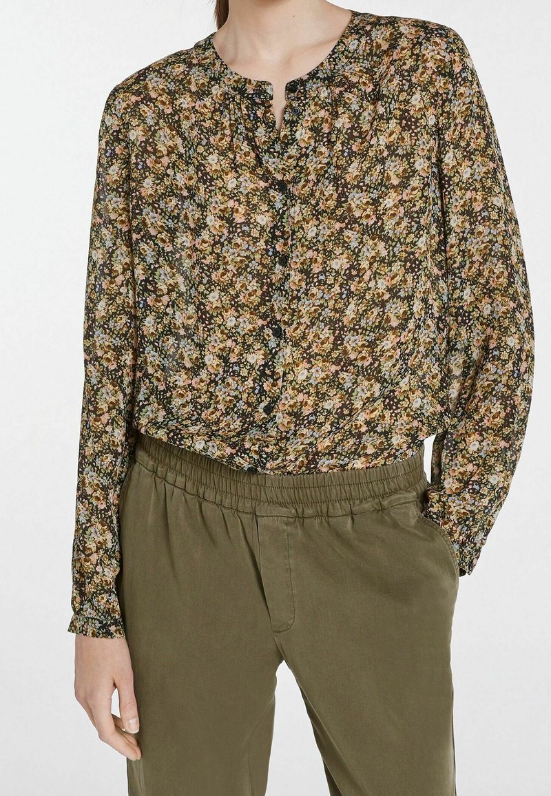 Blomstret bluse i mørkt stof med små pink og blå blomster, lange ærmer, en afslappet pasform og en knappet halsudskæring. Kombineret med olivengrønne bukser.