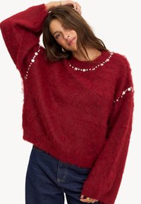 Pull en laine rouge flou, à coupe ample et manches larges, avec des accents circulaires en argent le long de l'encolure et des manches. Matériau texturé.