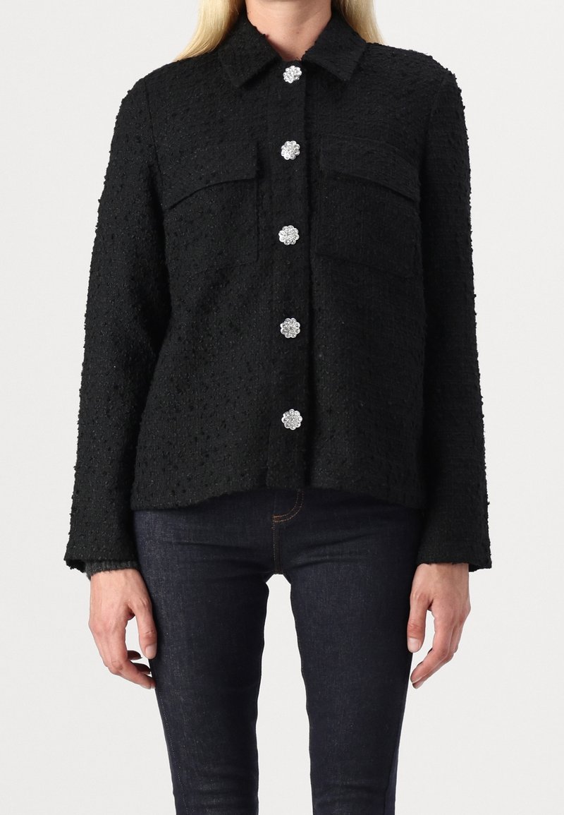 Femme portant une veste noire texturée à manches longues avec des boutons floraux argentés et un jean bleu foncé sur un fond uni.