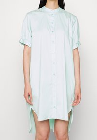 Robe chemise vert clair à manches courtes avec col montant, boutonnière à l'avant et poignets volants. Dispose d'échancrures sur les côtés pour un ajustement décontracté.