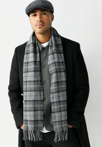 Man wearing grey plaid scarf, grey buttoned sweater, black overcoat, and grey flat cap, standing with hands in pockets on white background.