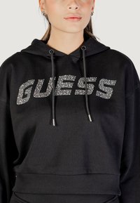 Haut à capuche noir avec "GUESS" en cristaux de rhinestones argentés, fermeture éclair avec cordon, et taille côtelée pour un look ajusté. Tissu doux et texturé.