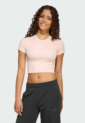 Světlé růžové sportovní tričko zkráceného střihu s krátkými rukávy a kulatým výstřihem, s malým bílým logem na hrudi. Kombinováno s tmavě šedými tepláky.