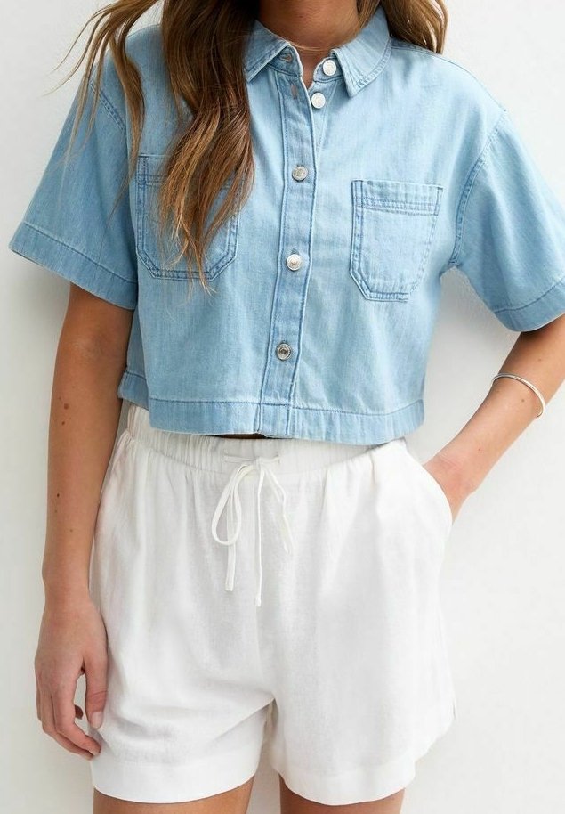 Chemise courte bleu clair à boutons avec deux poches poitrine, associée à un short blanc à cordon, portée par une personne debout avec une main dans la poche.