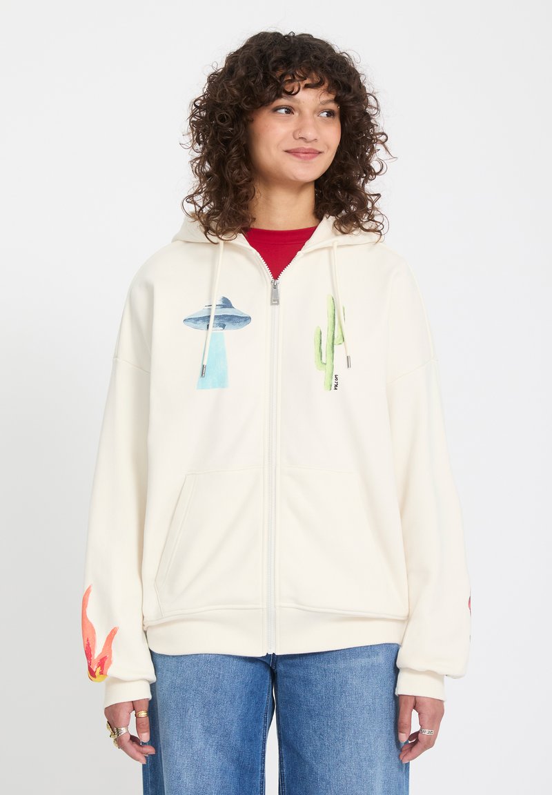 Jeune femme portant un sweat à capuche crème zippé avec des motifs de cactus et d'OVNI, assorti à un jean bleu, sur fond blanc uni.