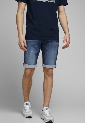 Mężczyzna w podwiniętych niebieskich szortach z denimu, czarnej koszulce z grafiką i białych butach Puma, stoi na szarym tle.