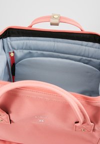 Růžová textilní taška se světle šedým vnitřkem, zapínáním na zip, dvojitými uchy a vnitřními přihrádkami. Má zlatý knoflík na zacvaknutí a logo.