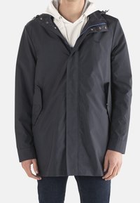 Dunkelblauer wasserdichter Herrenmantel mit Kapuze, Reißverschluss und Druckknöpfen, zwei seitlichen Taschen und glatter Textur. Über einem Hoodie getragen.