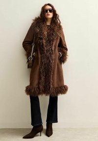 Bruine suede jas met nepbontafwerking, met een middellange ontwerp, brede kraag en zijzakken. Gepaard met donkere uitlopende spijkerbroek en hakken.