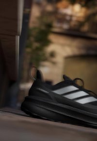 Svart Adidas Ultraboost löparsko med vita ränder som vilar inomhus på en träyta, med en suddig varm bakgrund.