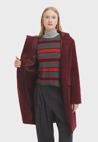 Burgunderfarbener Wollmantel mit einer plüschigen Textur, Knopfverschlüssen und einem satinierten Innenfutter, getragen über einem grau-rot gestreiften Pullover und gestreiften Hosen.