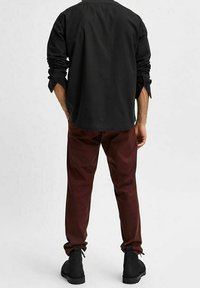 Hombre vistiendo una camiseta de manga larga negra, pantalones rojo oscuro y botas negras, de pie mirando hacia atrás contra un fondo claro.