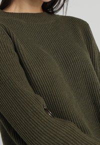 Frau trägt einen olivgrünen, geribbelten Strickpullover mit rundem Ausschnitt, der detaillierte Textur und einen kleinen Knopf am Ärmel zeigt.