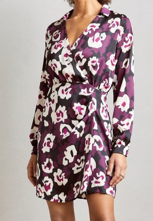 Frau trägt ein langärmeliges, Wickel-Minikleid mit einem abstrakten Blumenmuster in Lila, Schwarz und Weiß.