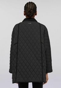 Person med flätat hår i knut, iklädd en svart vadderad jacka med diamantstygn, visad bakifrån mot en enfärgad bakgrund.