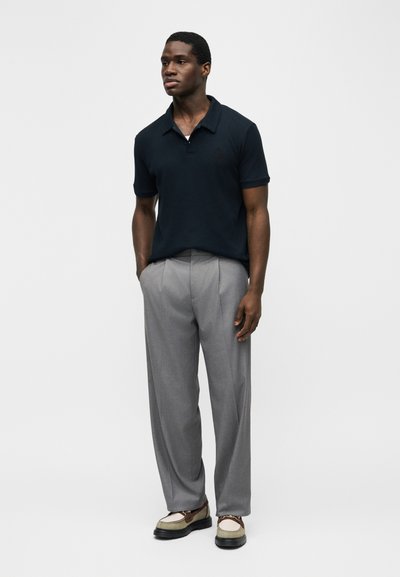 Homme debout, les mains dans les poches, portant un polo noir à manches courtes, un pantalon plissé gris et des mocassins bicolores sur fond blanc.