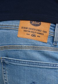 Lichtblauwe denim jeans met een tan leren label waarop "Petrol" staat en tekst over het vervaardigen van kwaliteitskleding. Beschikt over zichtbare stikseldetails.