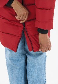 Röd vadderad jacka med dragkedja, texturerad yta och svarta innermuddar. Modellen har ljusblå denimjeans.