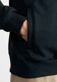 Sudadera negra con bolsillo tipo canguro, puños y dobladillo acanalados. Textura suave con un diseño sólido, vista de lado.
