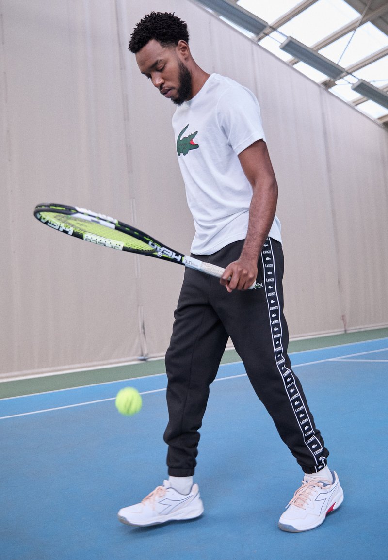 Tenisista w białej koszulce Lacoste i czarnych dresach z bocznym brandingiem, trzymający rakietę, przygotowujący się do uderzenia zielonej piłki tenisowej.