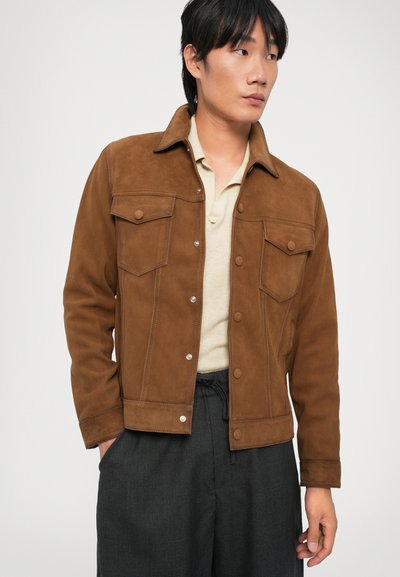 Braune Wildlederjacke mit Kragen, zwei Brusttaschen und silbernen Knöpfen; kombiniert mit einem beigen Poloshirt und dunkelgrauen Hosen.