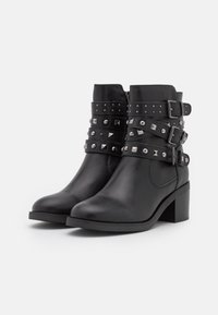 Botas de cuero negro hasta el tobillo con punta cerrada, que cuentan con múltiples correas decoradas con tachuelas plateadas y hebillas, y un tacón ancho.