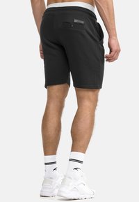 Ein Mann trägt schwarze Shorts mit einer geknöpften Gesäßtasche, weiße Sportsocken und weiße Turnschuhe und steht vor einem weißen Hintergrund.