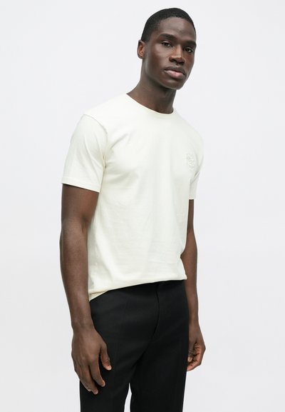 Camiseta de manga corta color crema claro con cuello redondo y logo en el pecho, combinada con pantalones negros. Hecha de tela de algodón suave.