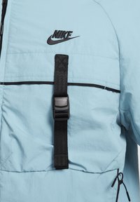 Ljusblå jacka i lättviktig tyg med en svart Nike-logga, justerbar svart rem med plastspänne och dragsko i nederkant.