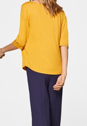 Kobieta ubrana w luźną, żółtą bluzkę z rękawami do łokcia i granatowe spodnie, pokazana od tyłu na jednolitym tle.