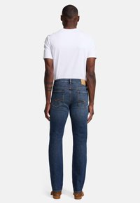 Jeans blu scuro con un design a gamba dritta, texture in denim, tasche posteriori e una patch in pelle beige in vita, abbinati a una t-shirt bianca.
