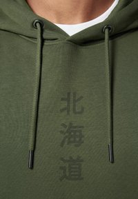 Haut à capuche vert olive avec une finition texturée, doté d'un cordon de serrage avec des accessoires noirs et d'un grand texte en japonais sur le devant.